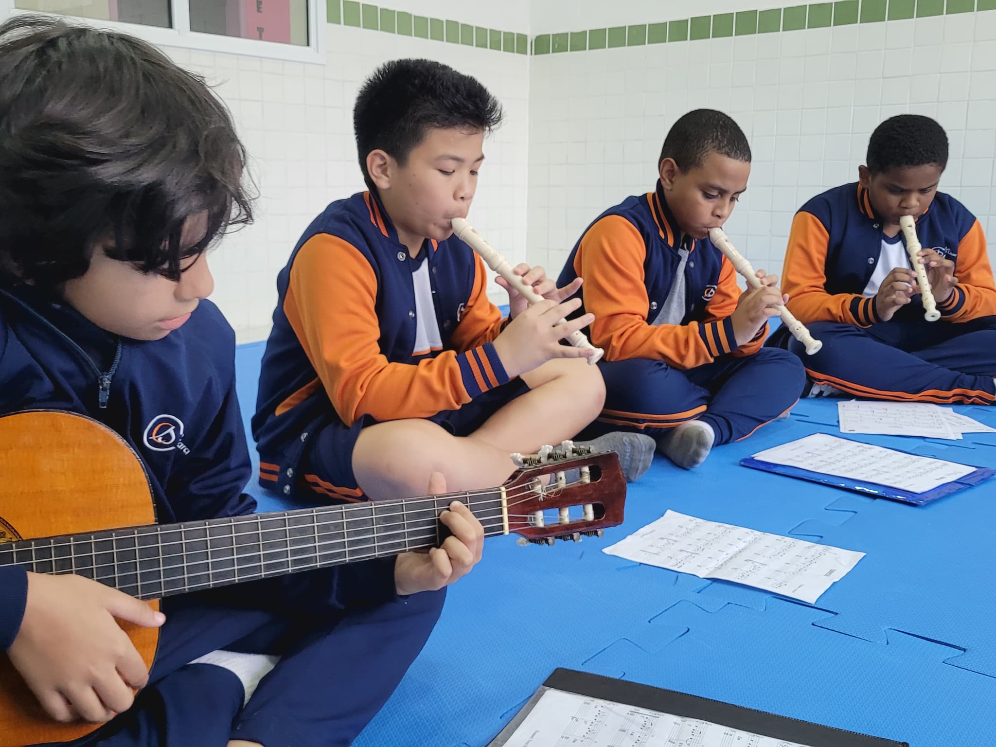 Estudantes participam de aula de música, tocando violão e flautas enquanto acompanham partituras.