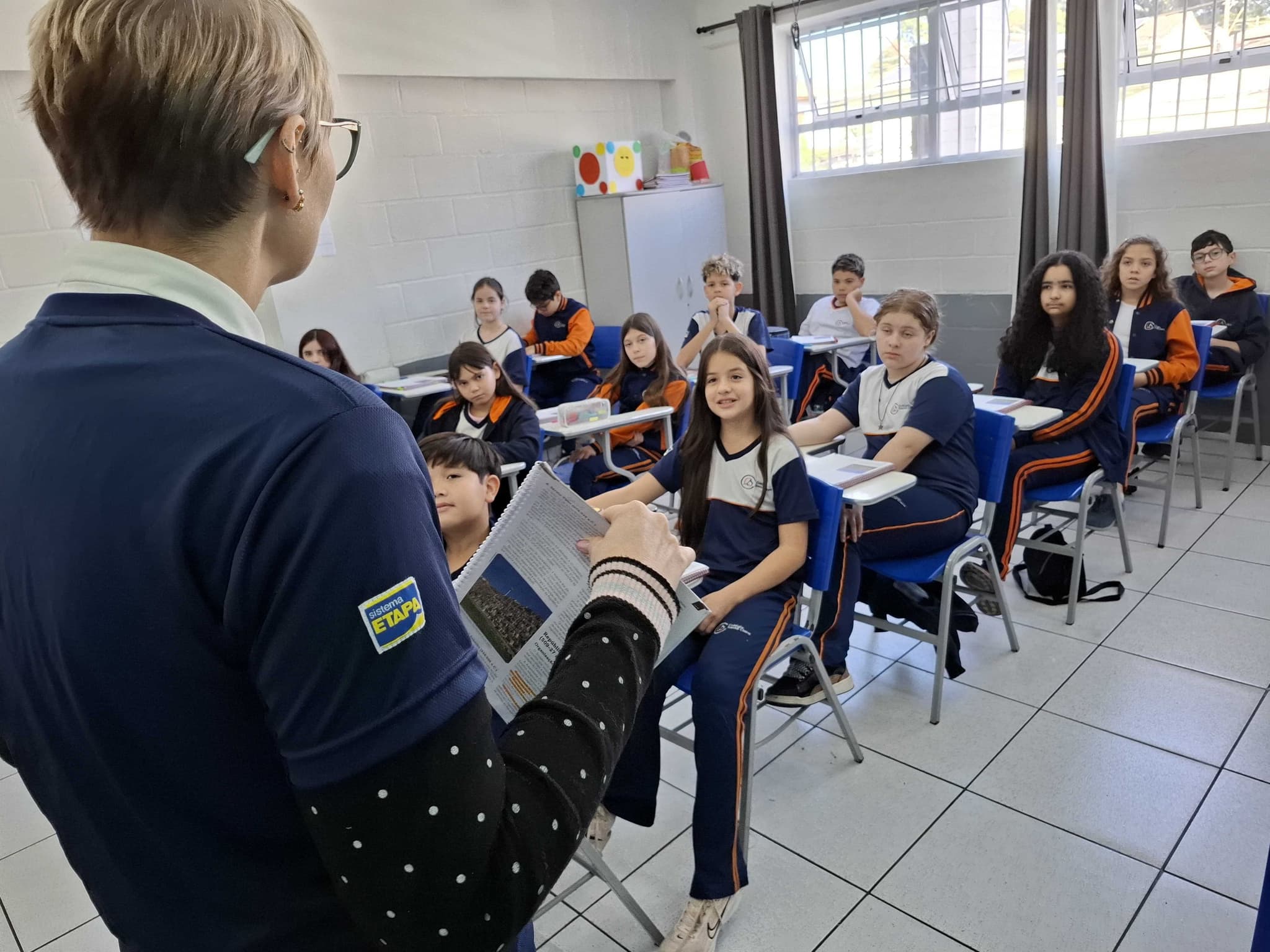 Professora em frente à turma durante aula, com estudantes sentados em suas carteiras e acompanhando a explicação.