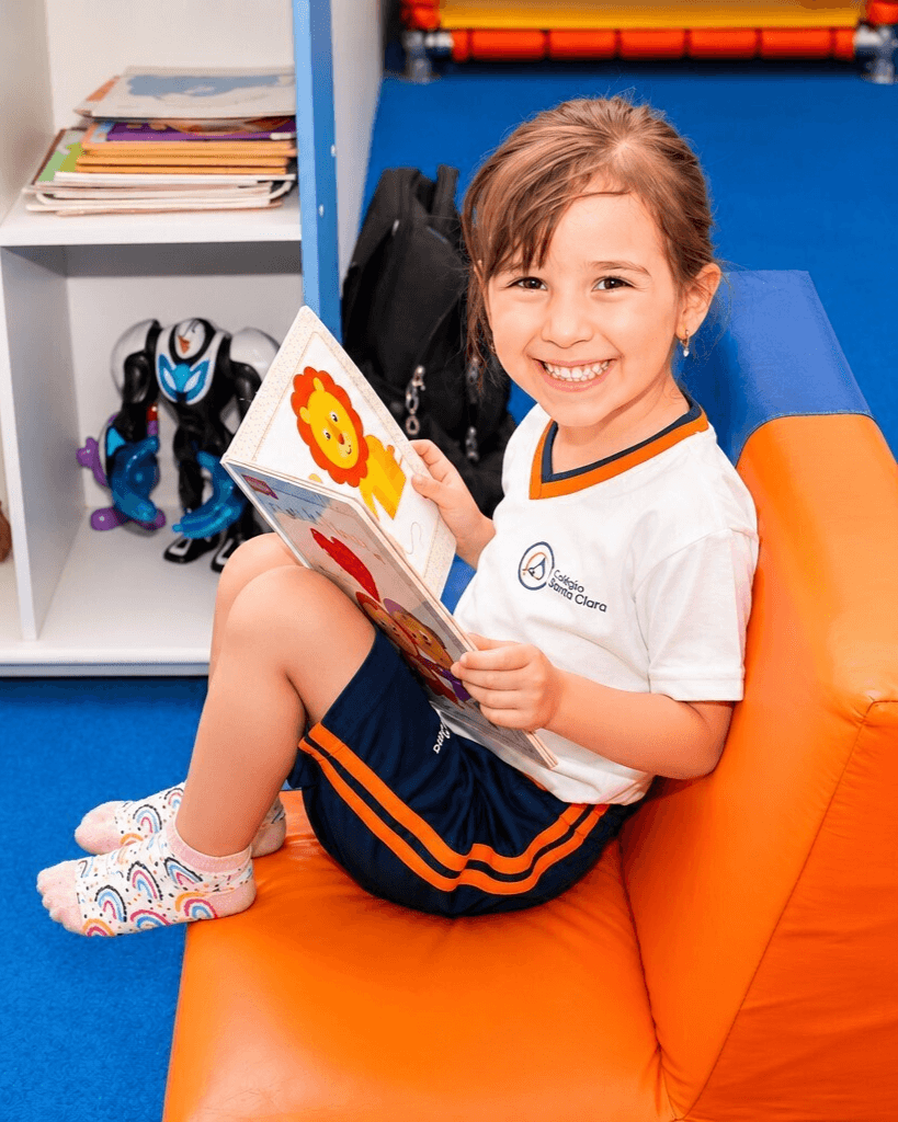Menina sorridente sentada em um sofá infantil, segurando um livro ilustrado.