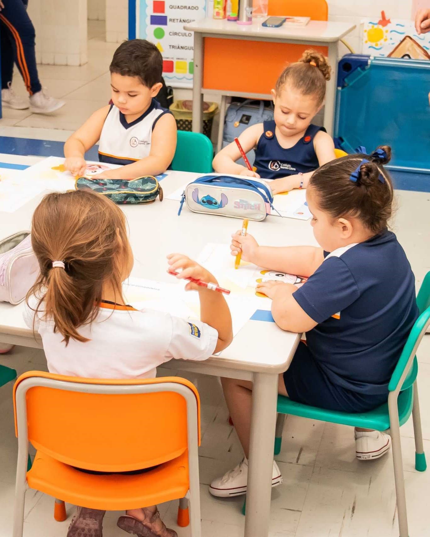 Crianças sentadas ao redor de uma mesa realizando uma atividade de desenho e escrita em sala de aula.