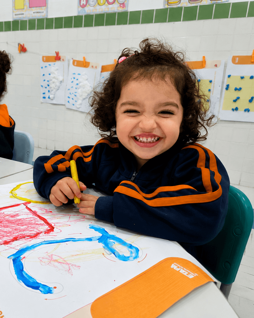 Menina sorridente desenhando com giz de cera durante atividade em sala de aula.