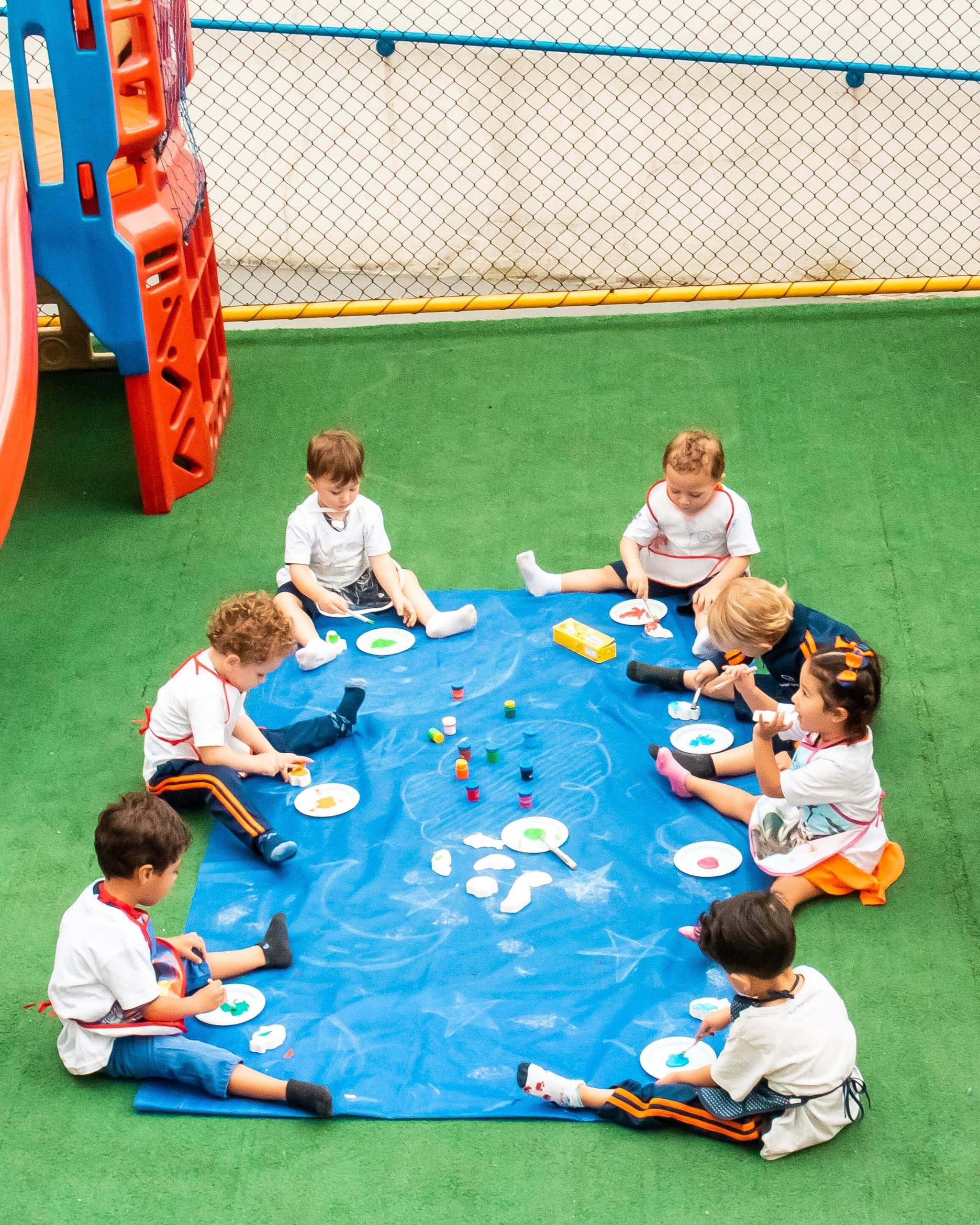 Grupo de crianças sentado em roda sobre uma lona azul, participando de uma atividade de pintura ao ar livre.