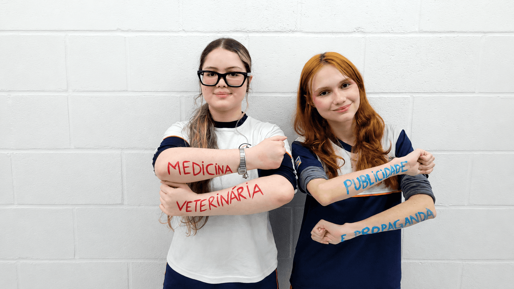 Duas estudantes posam lado a lado com nomes de cursos escritos nos braços, incluindo Medicina, Veterinária, Publicidade e Propaganda.