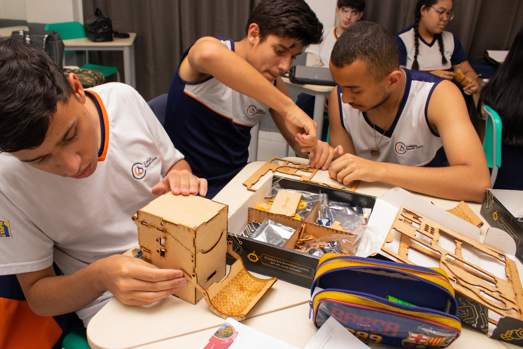 Estudantes em sala de aula montando kits de robótica e estruturas de madeira durante atividade prática em grupo.