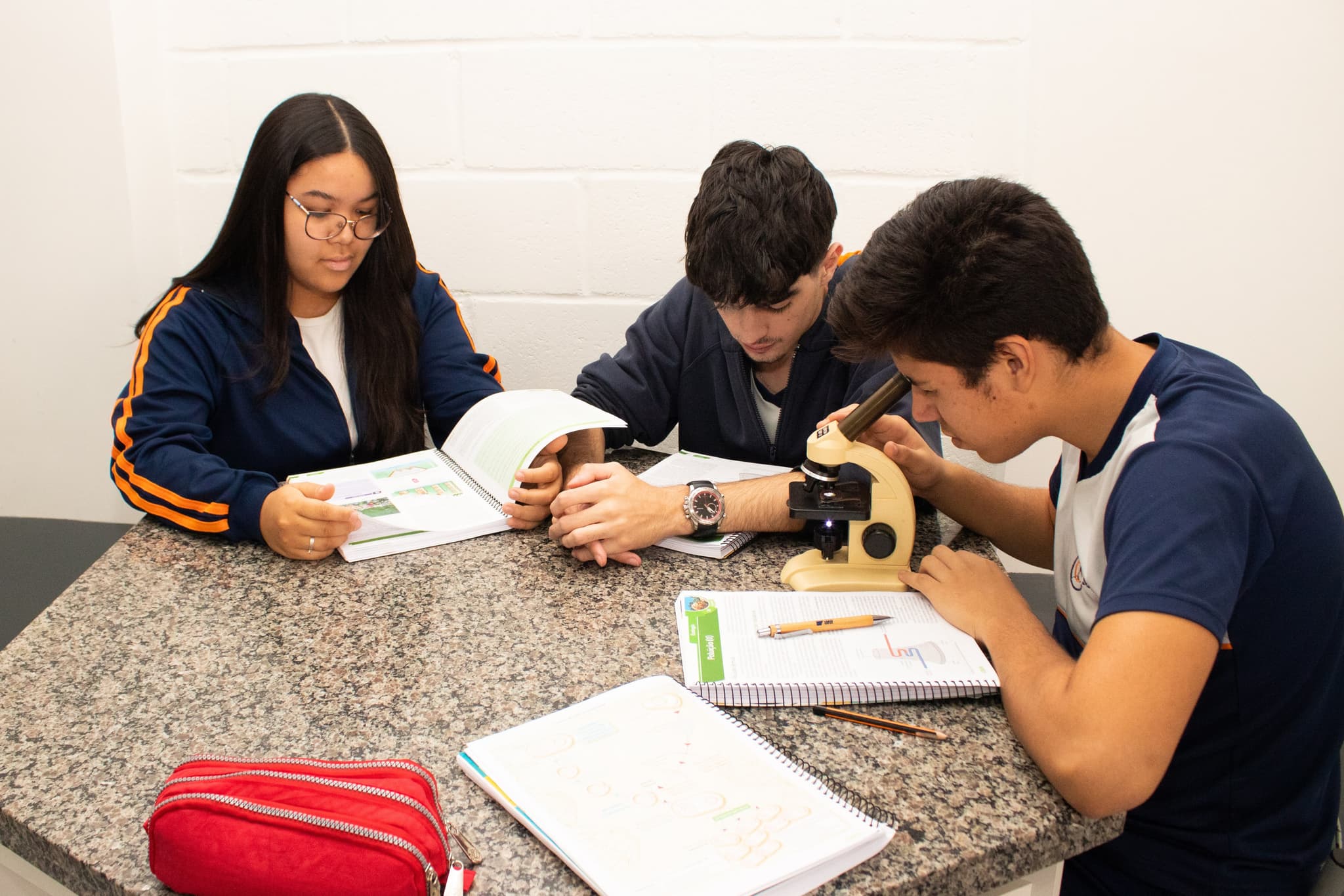 Três estudantes em laboratório escolar realizando atividade de ciências com microscópio e materiais didáticos sobre a bancada.